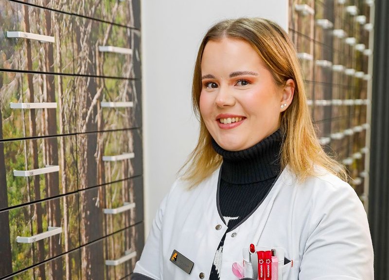 Nuori hymyilevä farmaseutti valkoisessa työasussa Kupittaan apteekissa. Hänellä on musta poolopaita takin alla, ja rintataskussa näkyy punaisia kyniä. Taustalla on lääkelaatikoita, joiden pinnassa on metsäaiheinen kuva.